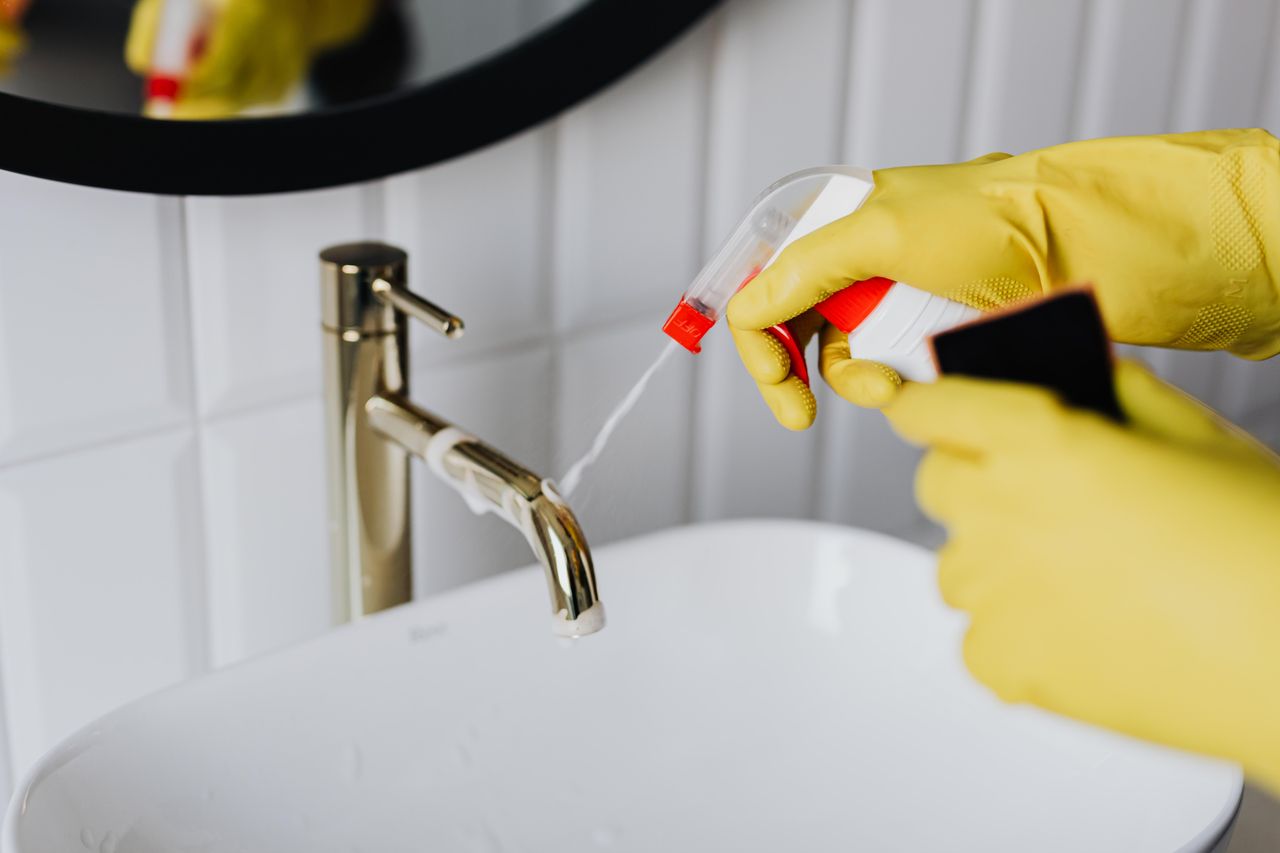 project-01-d From above, an anonymous housekeeper wearing yellow rubber gloves sprays cleaner onto a chrome bathroom water faucet